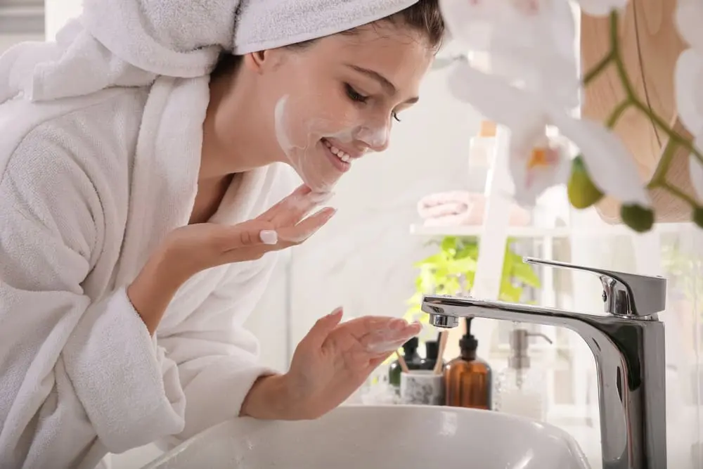 Person in a white bathrobe applies facial cleanser at a bathroom sink. Skincare items are on the counter.